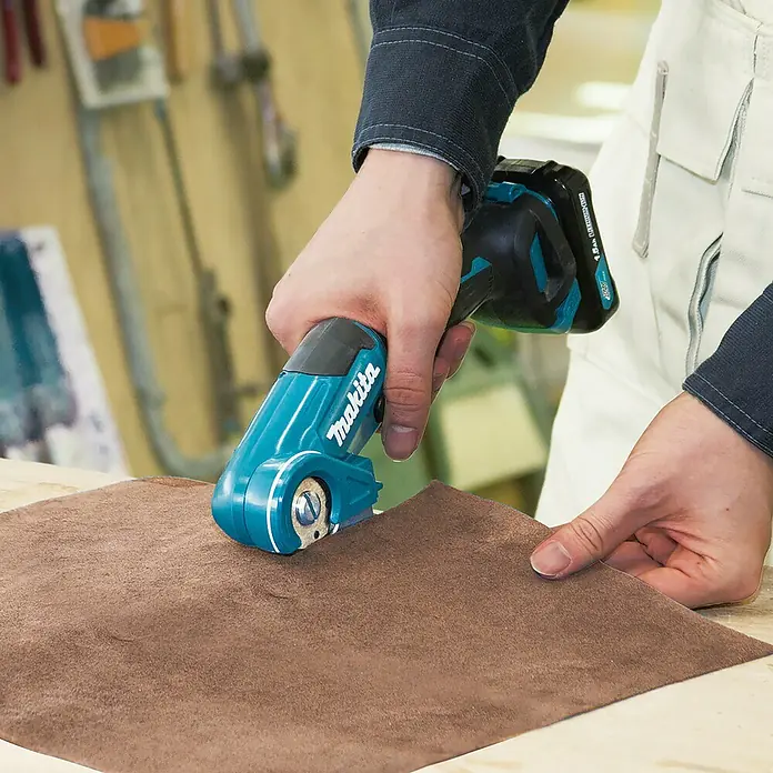 Makita CXT 12V max. Akku-Universalschere CP100DZ - 12 V, Ohne Akku, Schnitttiefe: Max. 6 mm - Person schneidet braunen Stoff mit einem türkisen Makita Akku-Rollenschneider.