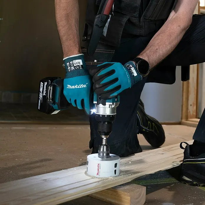 Makita LXT 18V Akku-Bohrschrauber DDF492RTJ - 18 V, 2 Akkus, 130 Nm - Eine Person mit Handschuhen verwendet einen Makita Bohrer mit weißem Lochsägeblatt, um ein Loch in ein Holzbrett zu schneiden.