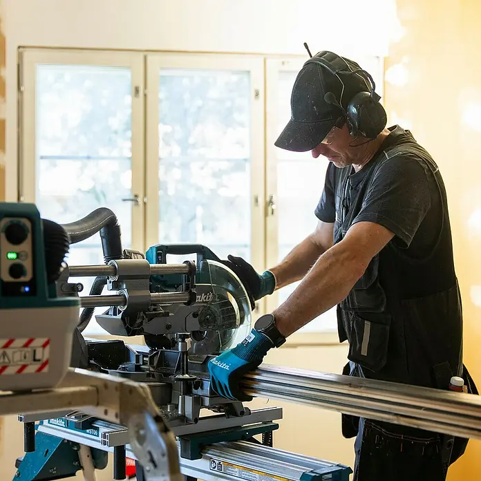 Makita XGT 40V max. Akku-Kapp- & Gehrungssäge LS002GZ01 - 40 V, Ohne Akku, 4.800 U/min - Eine Person schneidet ein silbernes Metallprofil mit einer türkis-schwarzen Gehrungssäge auf einem Ständer.