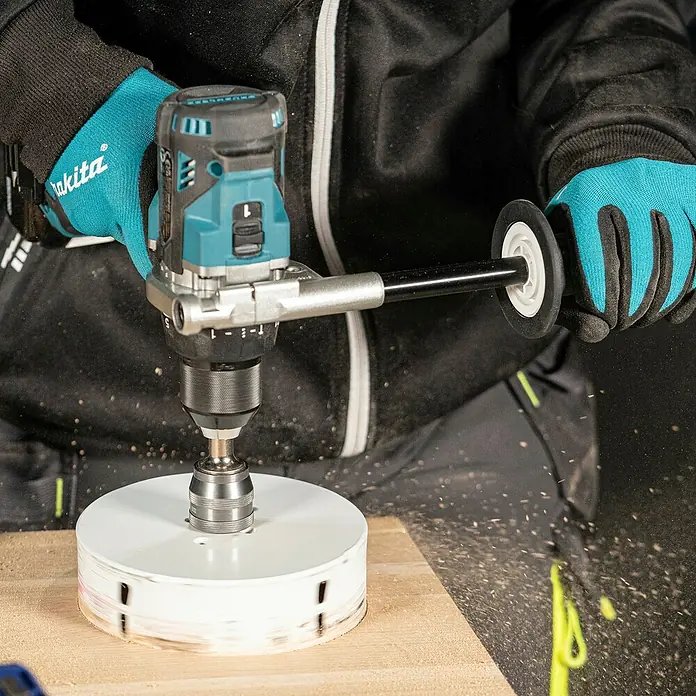 Makita LXT 18V Akku-Schlagbohrschrauber DHP492RTJ - 18 V, 2 Akkus, 5 Ah, 130 Nm - Person bohrt mit einer türkis-schwarzen Akku-Bohrmaschine und einer großen weißen Lochsäge ein Loch in ein Holzbrett.