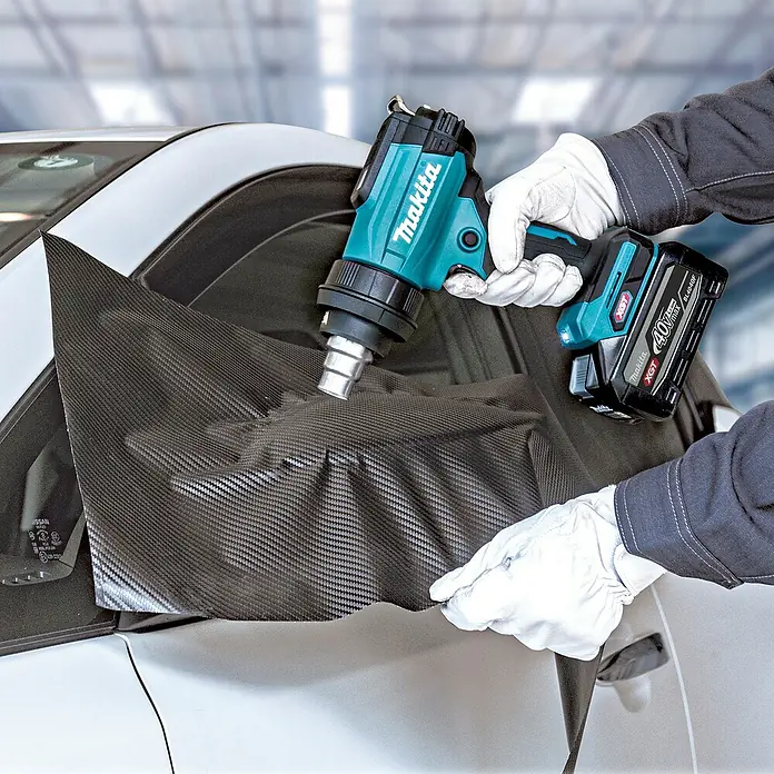 Makita XGT 40V max. Akku-Heißluftgebläse HG001G - 40 V, Ohne Akku, Temperaturbereich: 0 °C - 600 °C - Eine Person verwendet eine türkis-schwarze Makita Heißluftpistole mit 40V Akku, um eine Carbon-Autofolie auf ein weißes Auto aufzubringen.