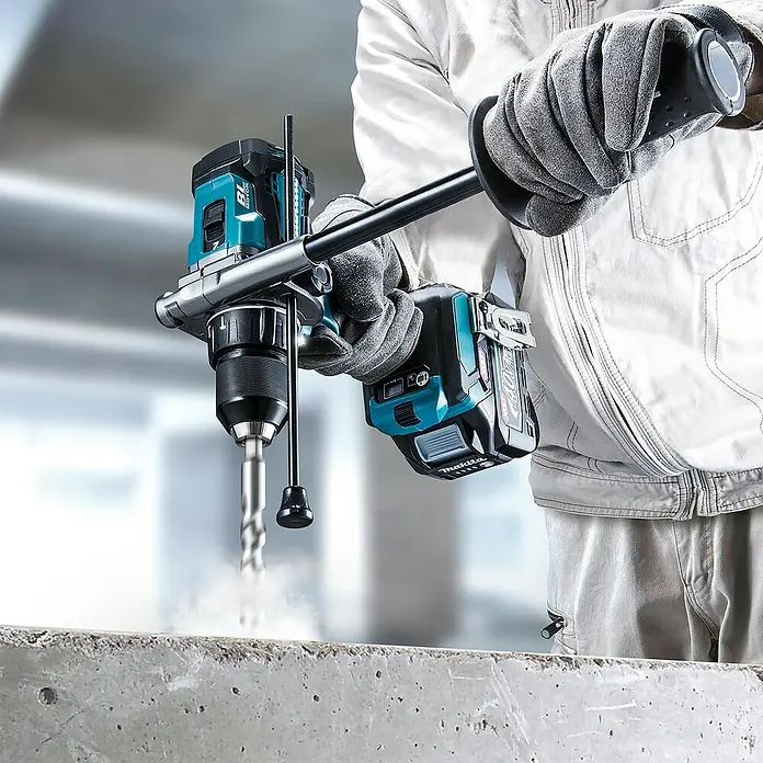 Makita XGT 40V max. Akku-Schlagbohrschrauber HP001GD201 - 40 V, 2 Akkus, 2,5 Ah, 41 Nm - Person in Arbeitshandschuhen bedient einen türkis-schwarzen Akku-Bohrhammer, der Staub erzeugt beim Bohren in Beton.