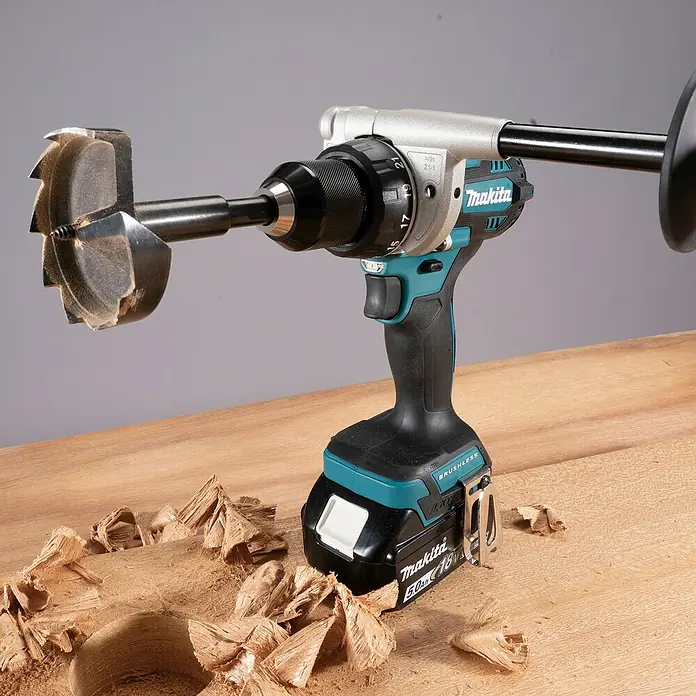 Makita LXT 18V Akku-Schlagbohrschrauber DHP492RTJ - 18 V, 2 Akkus, 5 Ah, 130 Nm - Türkis-schwarze Makita Akku-Bohrmaschine mit großem Holzbohrer und Zusatzhandgriff, bohrt in eine helle Holzoberfläche mit Spänen.