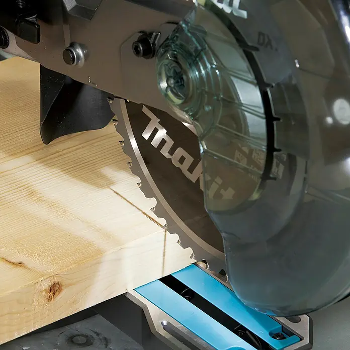 Makita XGT 40V max. Akku-Kapp- & Gehrungssäge LS002GZ01 - 40 V, Ohne Akku, 4.800 U/min - Nahaufnahme eines Makita Gehrungssägeblatts mit klarer Schutzhaube, das ein Holzbrett auf seiner blauen Basis schneidet.