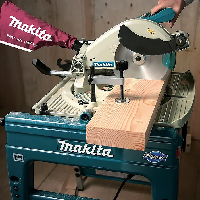Makita Tischkreissäge & Kapp-Gehrungssäge LF1000 - 1.650 W, Durchmesser Sägeblatt: 260 mm - Makita Gehrungssäge auf blaugrünem Ständer, schneidet ein Holzbrett. Cremefarbener Sägekopf und roter Staubfangsack sichtbar.