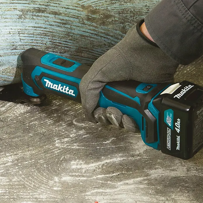 Makita CXT 12V max. Akku-Multifunktionswerkzeug TM30DZ - 12 V, Ohne Akku - Behandschuhte Hand bedient ein türkis-schwarzes Makita Multifunktionswerkzeug mit 12V 4.0Ah Akku, das Material vom Boden entfernt.