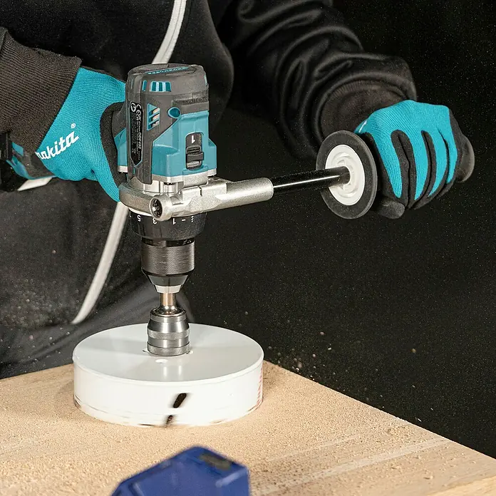 Makita LXT 18V Akku-Bohrschrauber DDF492RTJ - 18 V, 2 Akkus, 130 Nm - Eine Person bohrt mit einem türkis-schwarzen Makita Akku-Bohrschrauber und einer Lochsäge ein großes Loch in eine Holzplatte, wobei Sägemehl entsteht.