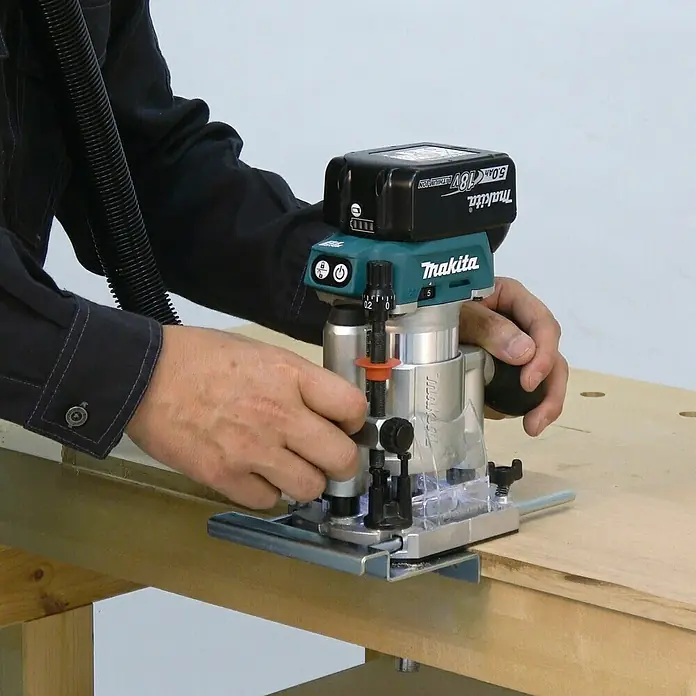 Makita LXT 18V Akku-Multifunktionsfräse DRT50ZJX2 - 18 V, Ohne Akku, Leerlaufdrehzahl: 10.000 U/min - 30.000 U/min - Eine Person führt eine silberne und türkisfarbene Makita 18V Akku-Oberfräse mit schwarzem Akku über ein Holzwerkstück auf einer Werkbank.