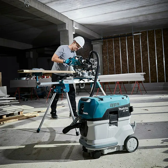 Makita XGT 40V max. Akku-Kapp- & Gehrungssäge LS002GZ01 - 40 V, Ohne Akku, 4.800 U/min - Türkis-weißer Makita Staubabsauger mit Schlauch auf Baustelle, vor Arbeiter mit Kappsäge.