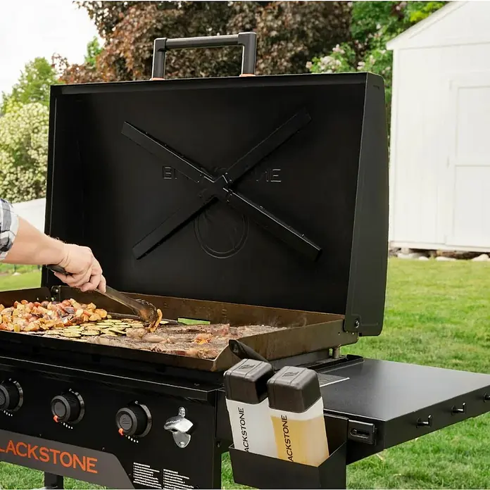 Blackstone Plancha Grill - Superficie de la parrilla (An x Pr): 91 x 55 cm, Negro, Número de quemadores: 4 ud. - Persona cocinando en una parrilla plana negra Blackstone con la tapa abierta. La parrilla cuenta con estantes laterales con botellas de condimentos.