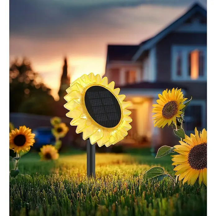 Globo LED-Solar-Dekoleuchte Sonnenblume - Lichtfarbe: Warmweiß, LED, Höhe: 29,5 cm - Eine leuchtende gelbe Solar-Gartenleuchte in Sonnenblumenform erhellt bei Dämmerung den Rasen, inmitten echter Sonnenblumen vor einem Haus.