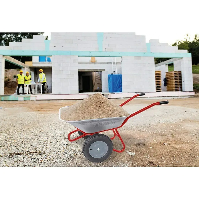 Gartenschubkarre "Zweirad" - 100 l, Stahl, Traglast: 160 kg - Eine silberne 'pro-bau-tec' Schubkarre mit rotem Gestell, gefüllt mit Sand, auf einer Baustelle. Unscharfe Bauarbeiter im Hintergrund.