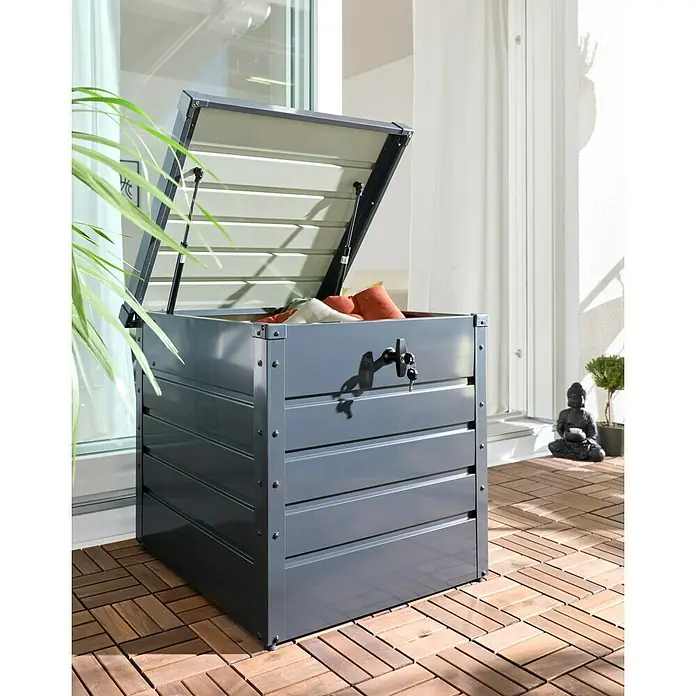 Hoggar Garten-Aufbewahrungsbox Sveg - L x B x H: 61 x 72 x 63 cm, Anthrazit, Metall - Dunkelgraue Outdoor-Staubox mit Lamellenstruktur, geöffnetem Deckel, bunten Kissen und Schloss auf Holzterrasse.