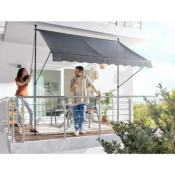 Sunfun Klemmmarkise / Senkrechtmarkise - Anthrazit, Breite: 3 m, Ausfall: 1,3 m - Dunkelgraue Balkonmarkise mit weißem Wellenrand spendet Schatten. Eine Frau bedient sie auf einem sonnigen Balkon.
