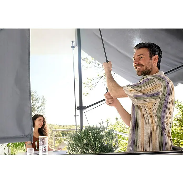 Sunfun Klemmmarkise / Senkrechtmarkise - Anthrazit, Breite: 3 m, Ausfall: 1,3 m - Mann stellt einen grauen Terrassenschirm ein, der eine Frau beim Lesen auf einem sonnigen Balkon beschattet.