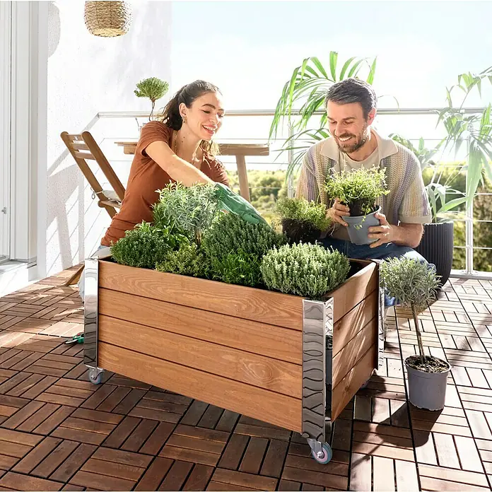 Dobar Hochbeet Rollfi - 78 x 52 x 40,5 cm, Holz, Braun - Hölzerner Pflanzkasten auf Rollen, gefüllt mit Kräutern, von einem Paar auf einem Balkon bepflanzt. Mit Naturholz und Metallakzenten.