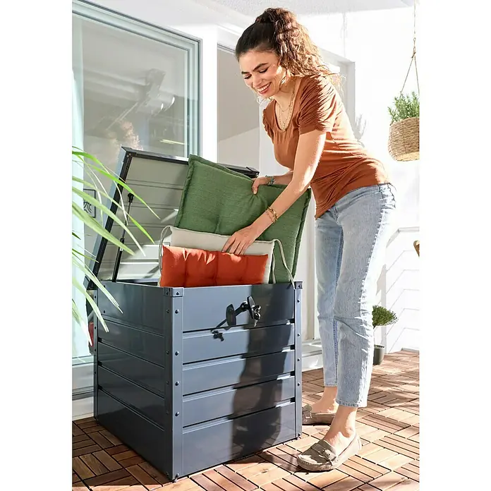 Hoggar Garten-Aufbewahrungsbox Sveg - L x B x H: 61 x 72 x 63 cm, Anthrazit, Metall - Dunkelgraue Outdoor-Aufbewahrungsbox auf einem Balkon, eine Frau legt grüne und orange Kissen hinein. Mit aufklappbarem Deckel und Verriegelungsmechanismus.