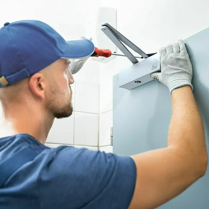 Basi Türschließer TS 200 - L x B x H: 210 x 40 x 50 mm, Silber - Mann installiert einen silber-grauen Türschließer mit einem Schraubendreher an einer Tür.
