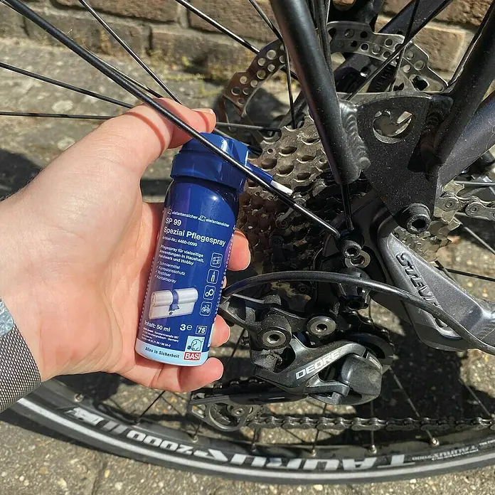 Basi Pflegespray - 50 ml - Hand hält blaue SP 99 Spezial Pflegespray-Flasche nahe dem Hinterrad und Schaltwerk eines Fahrrads.