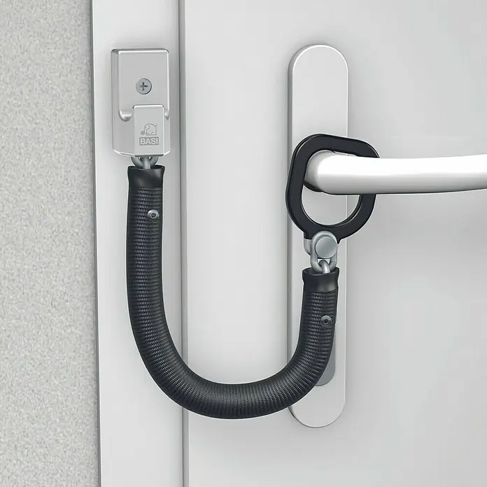 Basi Türkette TK 25 - Kettenlänge: 18,5 cm, Silber - Schwarze flexible Türsicherung mit silbernen Beschlägen, verbindet eine weiße Türklinke mit einer Wandhalterung mit BASI-Logo.