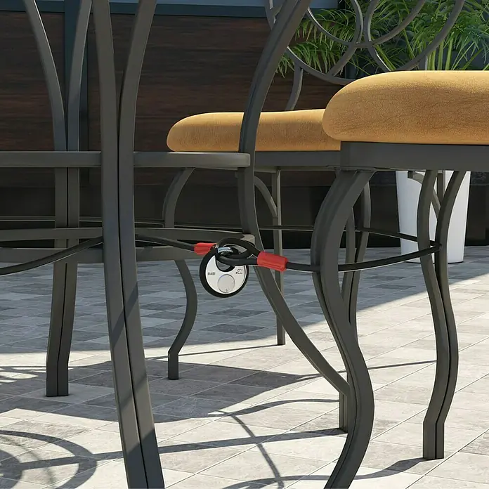 Basi Schlaufenseil SKA 500 - Ø x L: 10 mm x 5 m - Schwarzes Kabelschloss mit roten Akzenten, das zwei dunkle Terrassenstühle mit braunen Polstern auf einer Terrasse sichert.