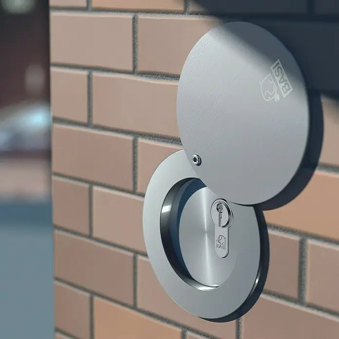 Basi Rohrtresor RT 103-S - Länge: 140 mm, Aluminium - Gebürstetes Sicherheitstürschloss aus Metall mit geöffneter Schutzabdeckung, Schlüsseloch und „BASI“-Logo, an einer Ziegelwand montiert.