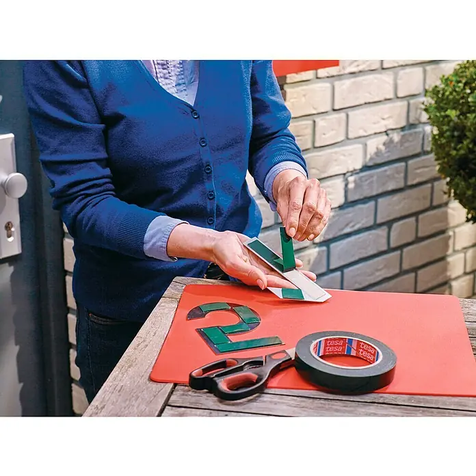 Tesa Powerbond Cinta de doble cara Ladrillo/Piedra - 1,9 cm x 5 m - Una persona pegando cinta de doble cara Tesa a números de casa sobre una esterilla roja, con tijeras y rollo de cinta cerca.