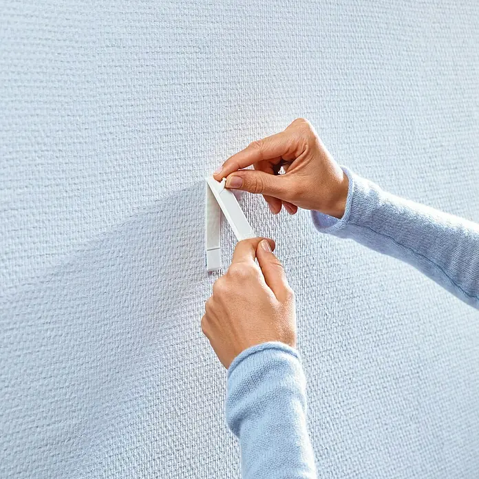 Tesa Clavo adhesivo - Apto para: Paredes pintadas, Carga soportada: 1 kg, 2 ud., Blanco - Una persona pega una tira adhesiva blanca a una pared con textura para colgar objetos.