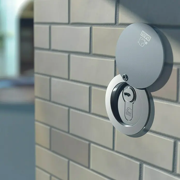 Basi Rohrtresor RT 103-S - Länge: 138 mm, Aluminium - Gebürsteter Metall-Schließzylinder mit offener drehbarer Abdeckung, die das Schlüsselloch enthüllt, an einer hellen Ziegelwand.