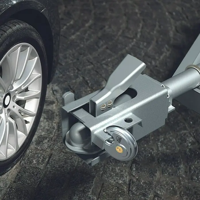 Basi Diskusschloss RVS 610 - B x H: 70 x 70 mm, Bügelstärke: 9,5 mm, Edelstahl - Metallisch silberne Anhängerkupplung mit Vorhängeschloss, an einem Anhängerarm neben einem Autorad auf Kopfsteinpflaster.