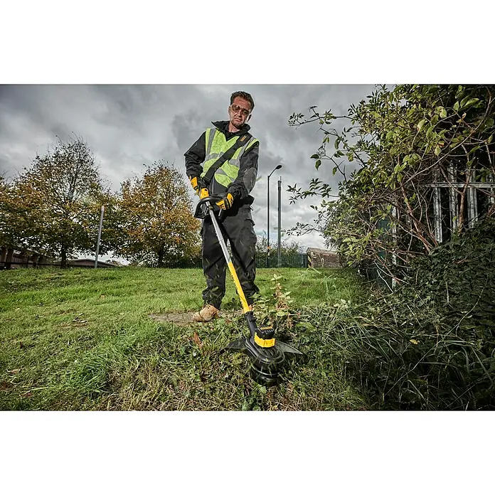 DeWalt XR 18V Akku-Rasentrimmer DCM561PBS-XJ - Ohne Akku, Schnittbreite Faden: 33 cm - Mann in Schutzkleidung benutzt einen gelb-schwarzen Rasentrimmer, um überwuchertes Gras zu entfernen.