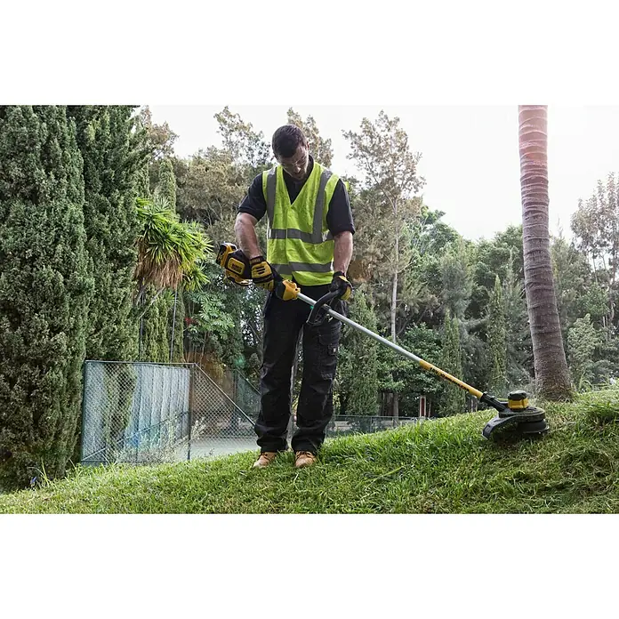 DeWalt XR 18V Akku-Rasentrimmer DCM561PBS-XJ - Ohne Akku, Schnittbreite Faden: 33 cm - Eine Person in Schutzkleidung verwendet einen gelb-schwarzen Rasentrimmer zum Schneiden von Gras auf einem Hang.
