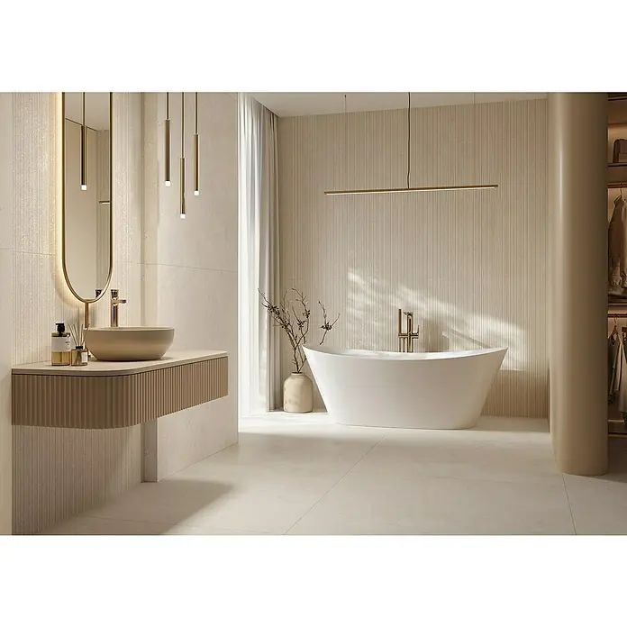 Wandfliese Manresa Lines - 39,8 x 119,8 cm, Beige, Matt - Ein modernes Badezimmer mit geriffelter beiger Waschtischplatte, Aufsatzwaschbecken, ovalem Spiegel, goldenen Pendelleuchten, freistehender Badewanne und strukturierten beigen Wänden.