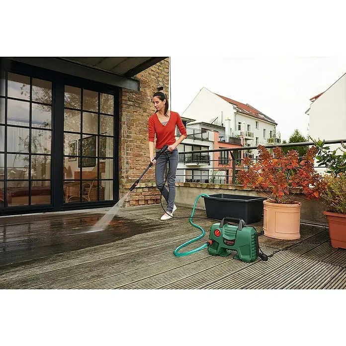 Bosch Hochdruckreiniger EasyAquatak 110 - 1.300 W, Max. Druck: 110 bar, Fördermenge: 330 l/h - Eine Frau reinigt mit einem grün-schwarzen Bosch EasyAquatak 110 Hochdruckreiniger eine Holzterrasse.