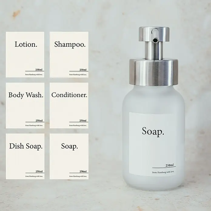 Designfabrik Hamburg Seifenspender - Edelstahl, Silber/Satiniertes Glas - Mattglas-Spender mit Edelstahlpumpe, gezeigt mit cremefarbenen Etiketten für Seife, Shampoo und andere Bad-/Küchenflüssigkeiten.