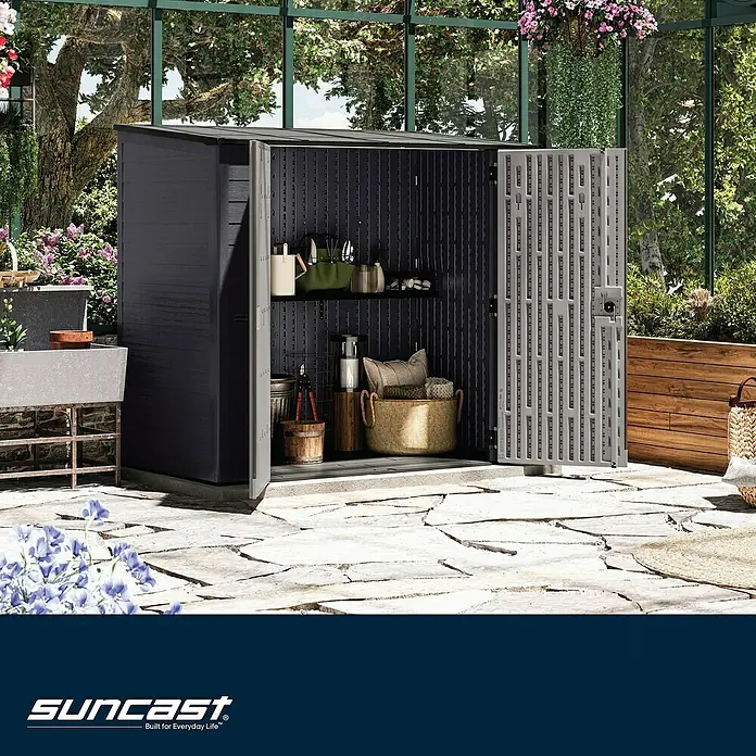 Suncast Geräteschrank - Kunststoff - Dunkelgrauer Geräteschuppen aus Kunstharz mit offenen Türen, der organisierte Regale und Gegenstände zeigt, auf einer Steinterrasse in einem Wintergarten.