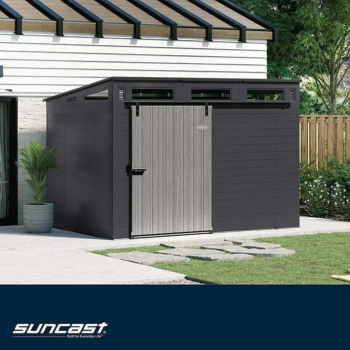 Suncast Gerätehaus - Kunststoff - Dunkelgrauer Gartenschuppen mit heller Schiebetür in Holzoptik, im Garten neben einem Haus.