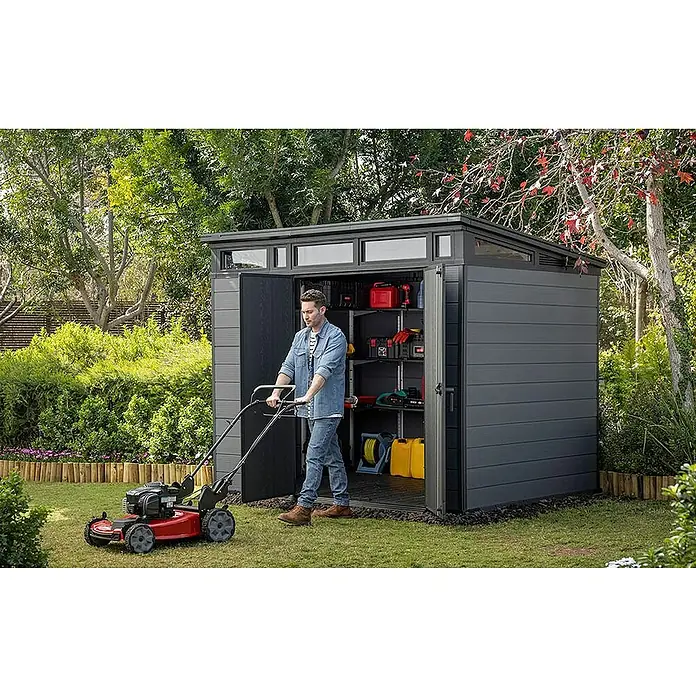 Keter Caseta de jardín Cortina 7x9 - Medidas ext. (An x Pr): 279 x 218 cm, Plástico, Gris, 6,08 m² - Caseta de jardín gris oscuro con herramientas organizadas, un hombre cortando el césped.