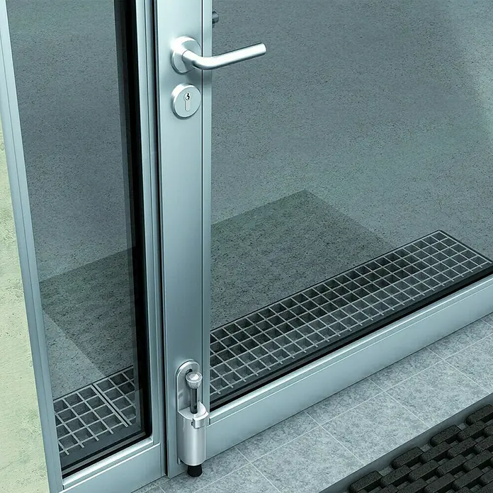 Basi Türstopper TFS 25 - B x H: 29 x 157 mm - Silberner Metall-Türstopper hält eine moderne Glastür mit Griff und Schloss offen; Ablaufrost sichtbar.