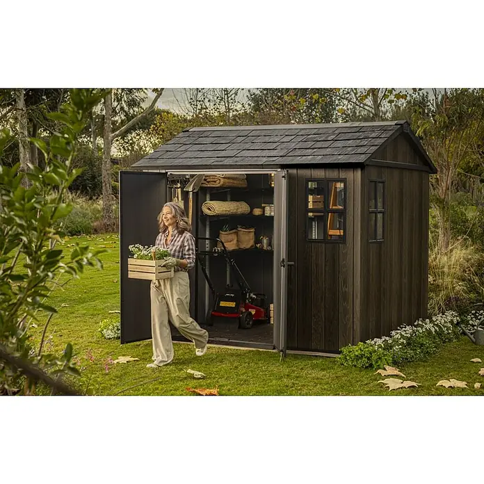 Keter Caseta de jardín Apex 759-975 - Medidas ext. (An x Pr): 287 x 230 cm, Plástico, Nogal/Negro - Caseta de jardín oscura efecto madera con techo de tejas, almacena herramientas. Una mujer sale con flores.