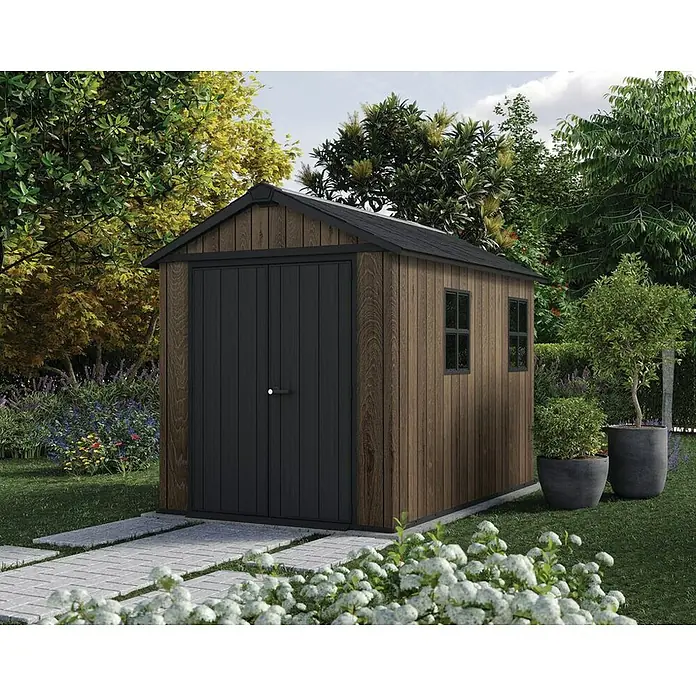 Keter Caseta de jardín Apex 759-975 - Medidas ext. (An x Pr): 287 x 230 cm, Plástico, Nogal/Negro - Caseta de jardín marrón con aspecto de madera, puertas y techo oscuros, en un jardín exuberante con sendero pavimentado.