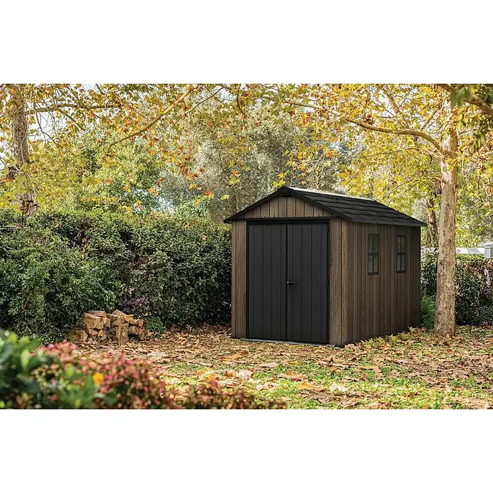 Keter Caseta de jardín Apex 7511-1175 - Medidas ext. (An x Pr): 350 x 230 cm, Plástico, Nogal/Negro - Caseta de jardín marrón con aspecto de madera, puertas dobles negras y techo, ubicada entre árboles y arbustos en un jardín natural.