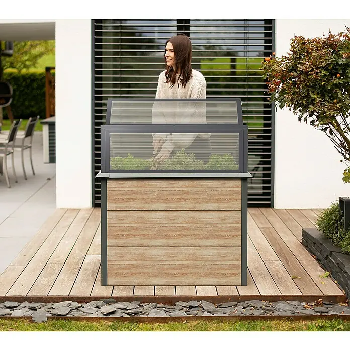 Westmann Hochbeet Primus Multi Plus M mit Frühbeetaufsatz - 50 x 100 x 113 cm, Metall, Braun/Anthrazit - Hochbeet/Frühbeet mit Holzoptik-Basis und grauem Rahmen, transparentem Deckel. Eine Frau pflegt darin Pflanzen auf einer Terrasse.