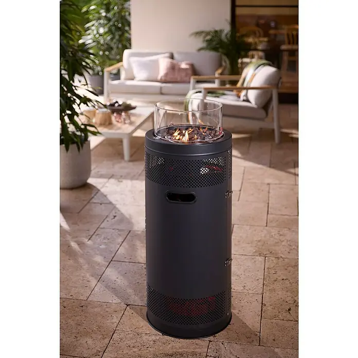 Kingstone Feuerstelle Sunset - Ø x H: 38 x 99 cm, Schwarz, Leistung: 5 kW - 9 kW - Dunkelgraue zylindrische Outdoor-Feuerstelle mit Glaswindschutz, Flammen und Lavasteinen, auf einer Terrasse.
