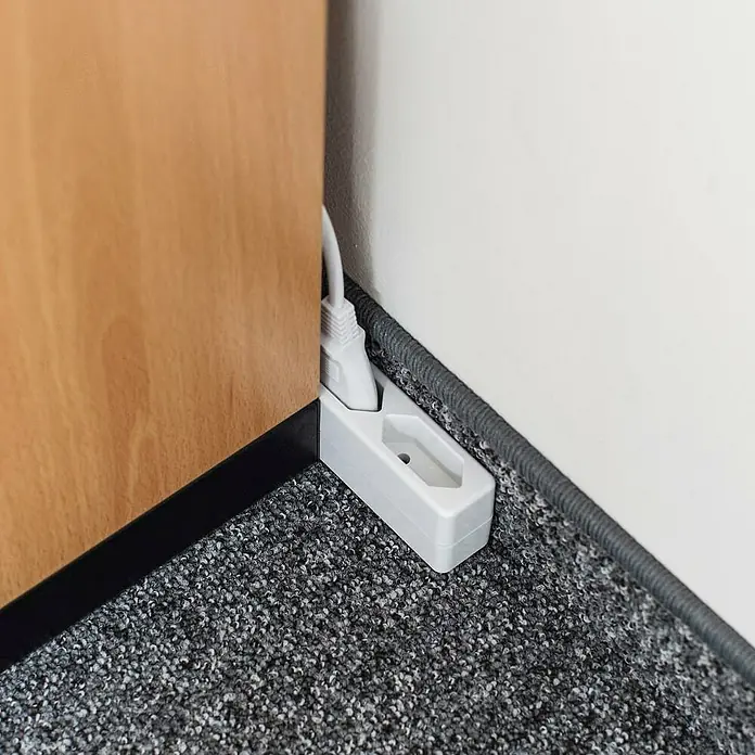 Hama Steckdosenleiste 3-fach 223020 - Kabellänge: 2,5 m - Weiße, flache Steckdosenleiste auf grauem Teppich, teilweise von Holzmöbeln verdeckt, ein Stecker eingesteckt.