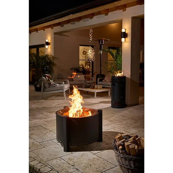 Kingstone Feuerschale Clear Flame - Durchmesser: 70 cm, Stahl - Schwarze Metall-Feuerstelle mit loderndem Feuer, auf einer Terrasse mit einem Korb Brennholz.