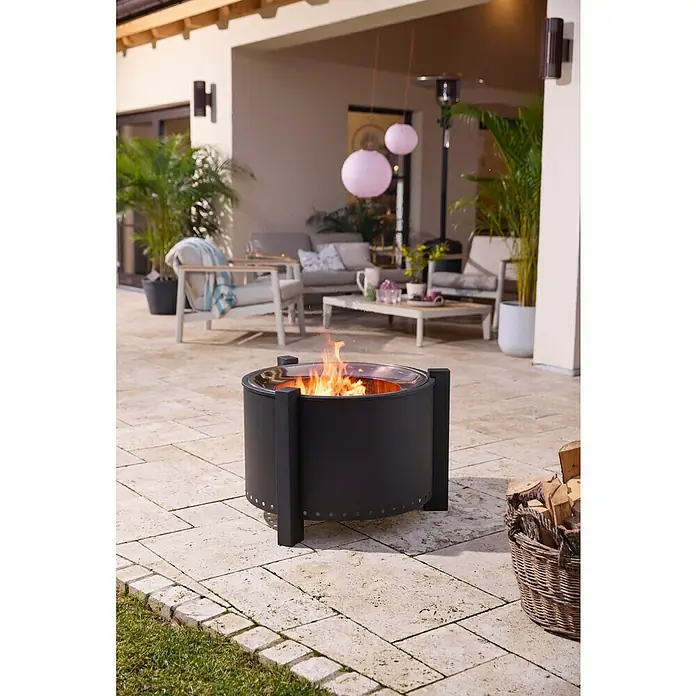 Kingstone Feuerschale Clear Flame - Durchmesser: 70 cm, Stahl - Schwarze Metall-Feuerschale mit Flammen auf einer hellen Terrasse, neben einem Korb Brennholz.