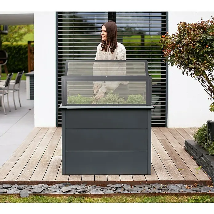 Westmann Hochbeet Primus Plus M mit Frühbeetaufsatz - 50 x 100 x 113 cm, Metall, Anthrazit - Dunkelgraues Hochbeet mit transparenter Abdeckung, eine Frau pflegt Pflanzen darin auf einer Holzterrasse.