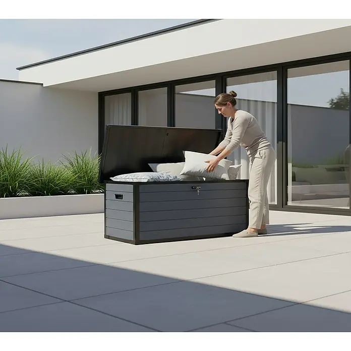 Westmann Kissenbox Silva - 74 x 150 x 66 cm, Kunststoff, Grau - Dunkelgraue Outdoor-Aufbewahrungsbox mit holzähnlicher Lamellenstruktur, geöffnet auf einer Terrasse, in die eine Frau Kissen legt.