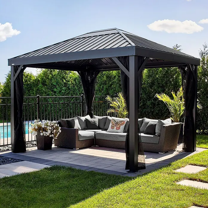 Sojag Standard-Pavillon Kiruna 1012 - Anthrazit, Metall, L x B x H: 299,3 x 364,3 x 297,6 cm - Dunkelgrauer Metallpavillon mit schwarzen Netzvorhängen, der ein geflochtenes Outdoor-Sofa mit grauen Kissen und einem Schmetterlingskissen bedeckt.
